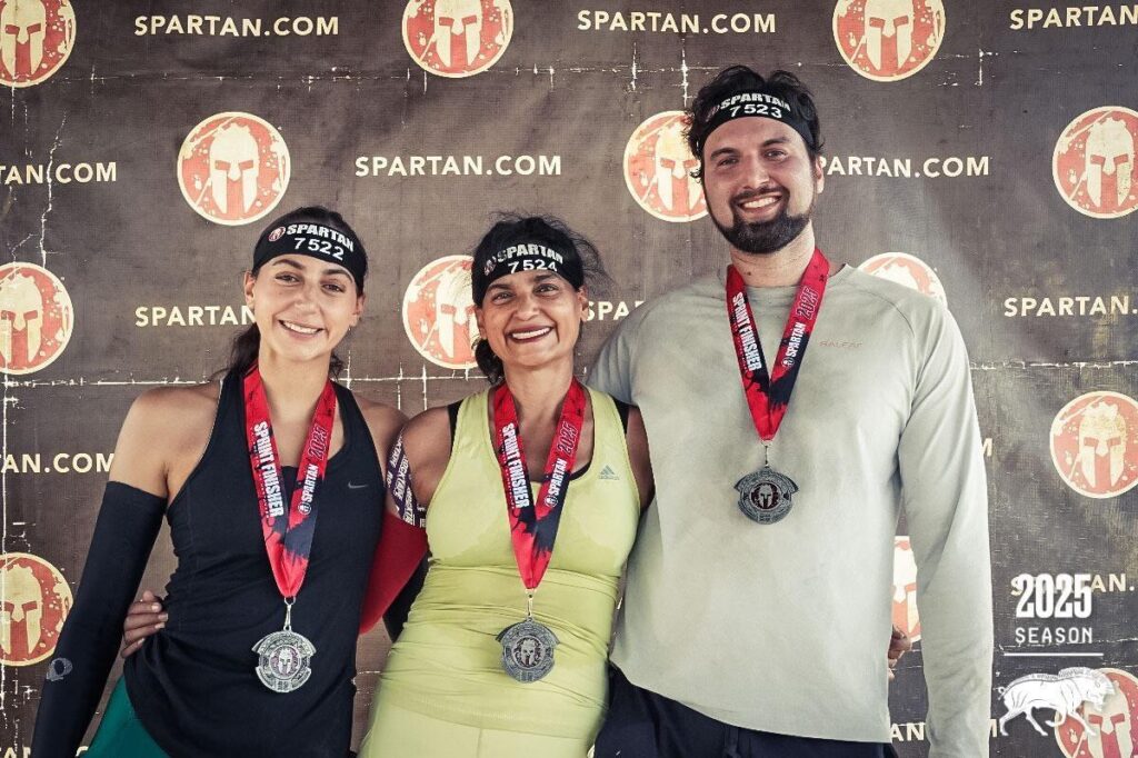 Three Spartan Race finishers wearing medals and headbands stand smiling in front of a Spartan.com backdrop, celebrating their achievement.