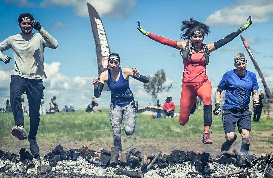Four Spartan Race participants leap over burning logs during an outdoor obstacle course, showcasing teamwork, endurance, and determination.