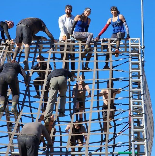 Spartan Race athletes climb a tall cargo net obstacle under a clear blue sky, demonstrating strength, balance, and camaraderie.